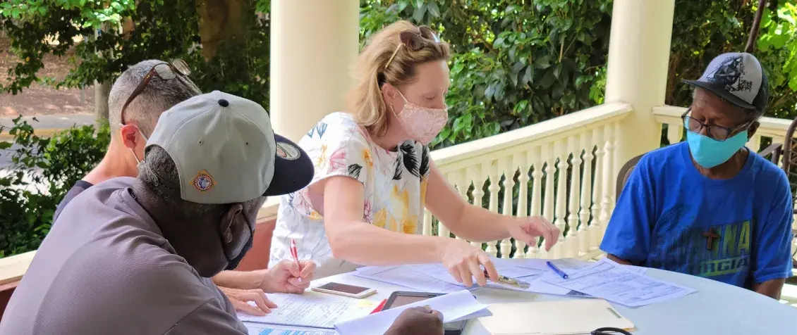A woman and three men, all wearing masks, gather around a table with paperwork