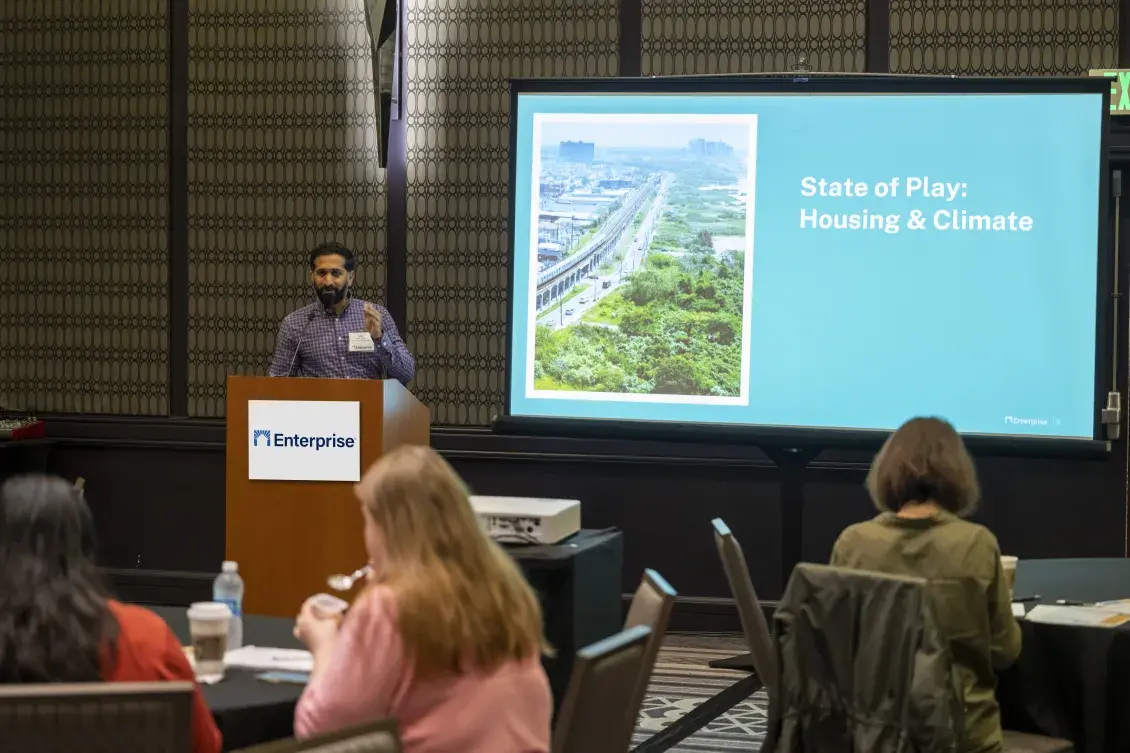 Speaker stands at a podium speaking to an audience in a conference room. To the speaker's right is a screen with a photo showing an aerial view of a city and the text: State of Play: Housing & Climage