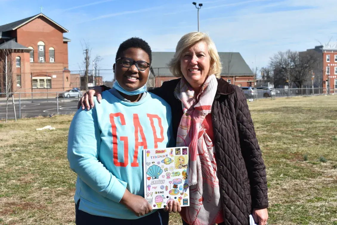 A young mentee and mentor stand at a park with apartment buildings in the background 