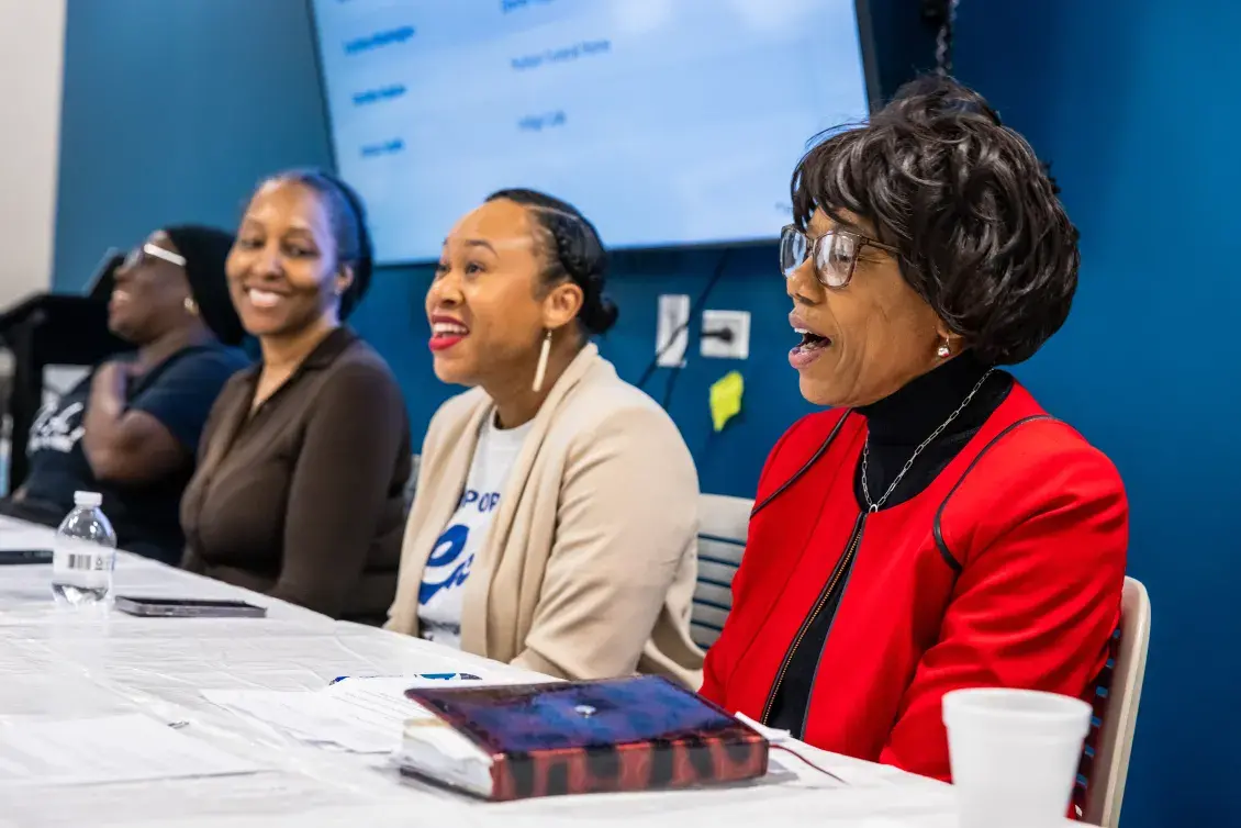 Four people sitting next to each other at a long table. In the background is a blue wall and video monitor.