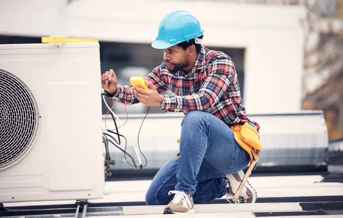 Person in plaid shirt wearing a blue construction hat kneels down to work on a heat pump.