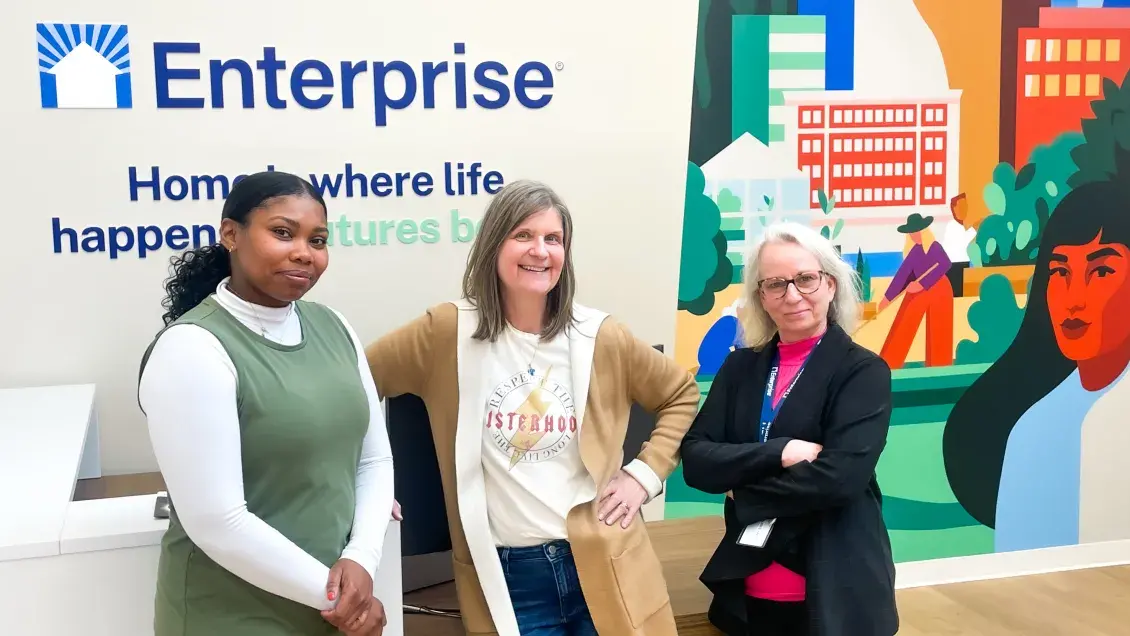 Three people stand in front of a wall imprinted with Enterprise, a quote, and a mural 
