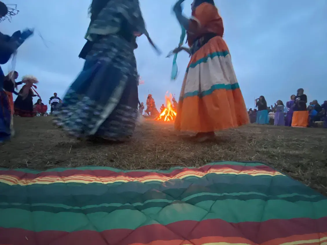 People wearing colorful skirts dance on a field with a bonfire burning in the background and a colorful blanket in the foreground
