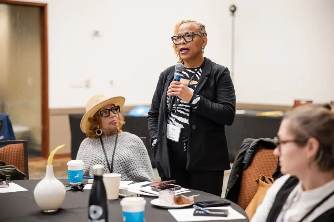 A woman stands with a microphone, and two people are seated on each side of her
