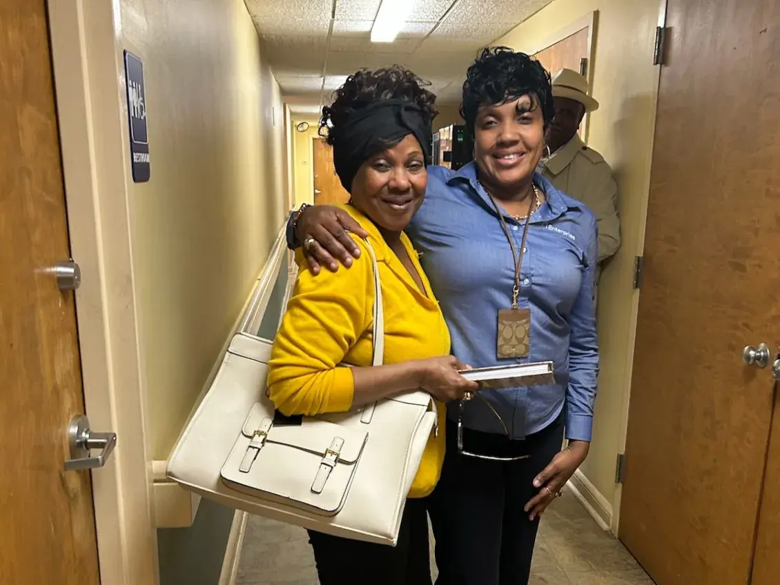 Two people stand next to each in a hallway, with the person on the right wearing a blue shirt and wrapping her arm around the shoulder of the person in a yellow shirt who carries a large white tote purse.