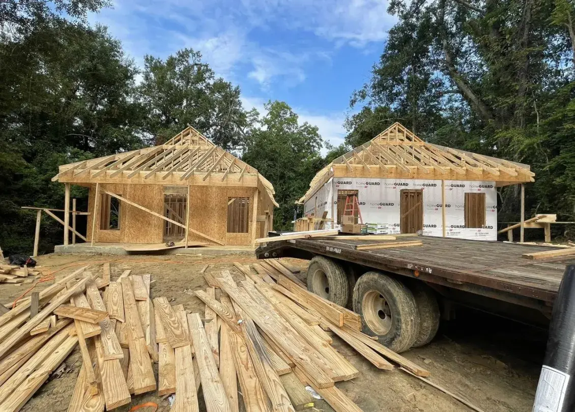 Two homes under construction stand side by side with a blue sky and treetops in the background