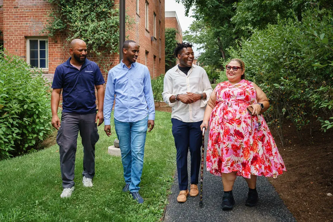 Group of four people walking down a path with trees and a brick building in the background