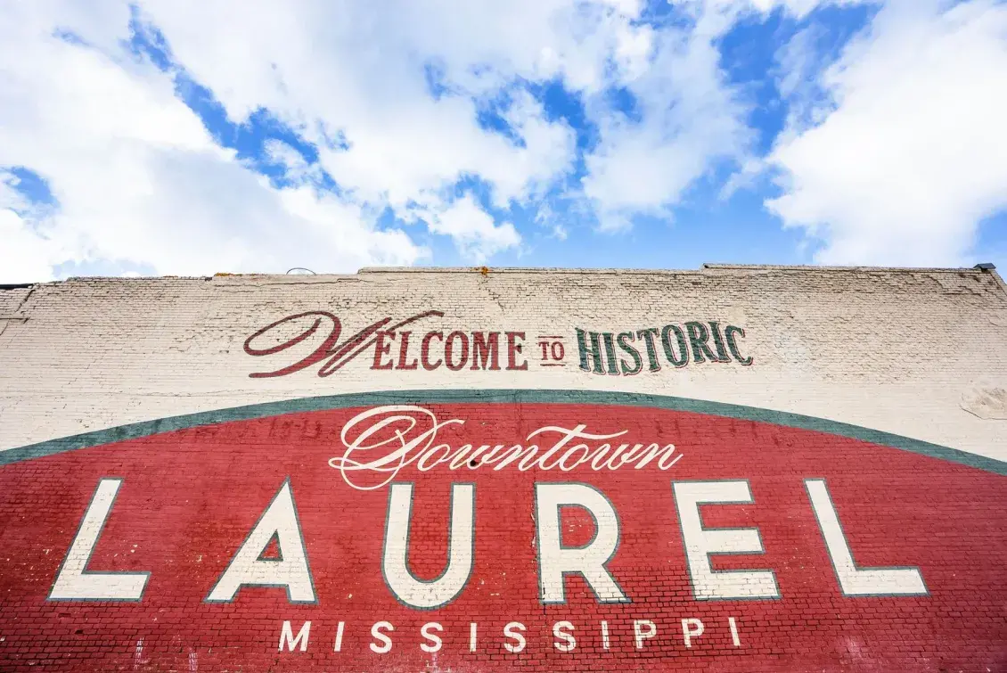 Red sign painted on a brick wall that reads: Welcome to Historic Downtown Laurel Mississippi, with a bright blue sky and white puffy clouds in the background.