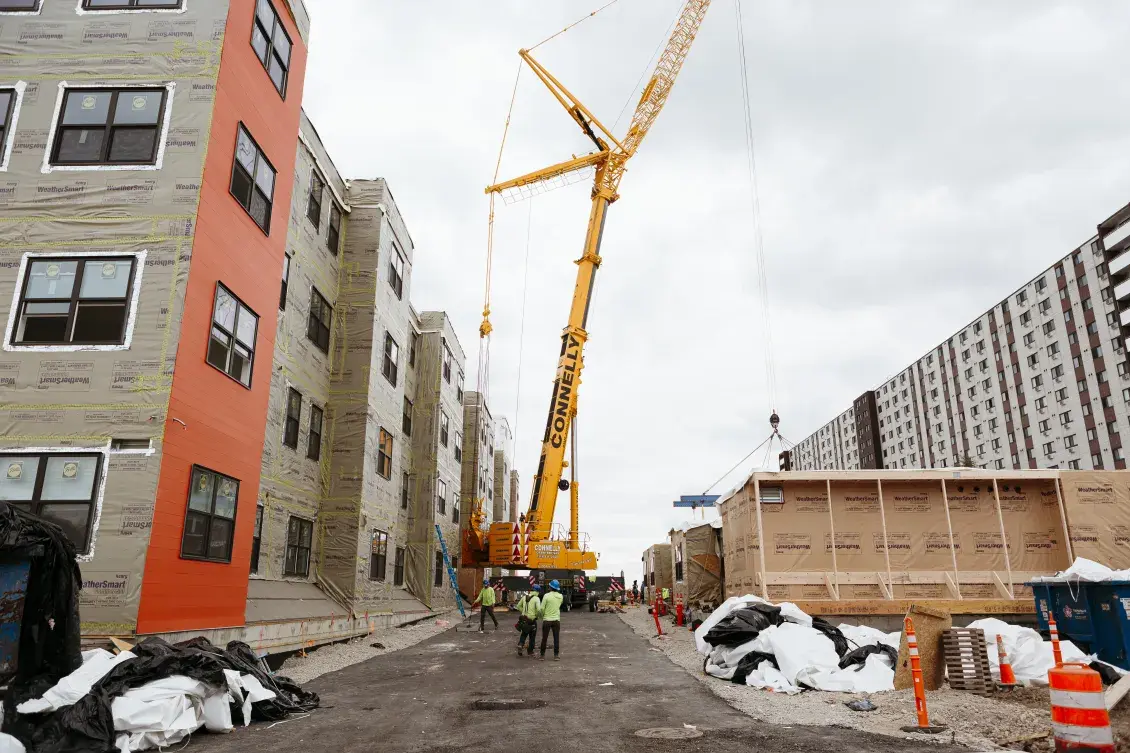 A crane sits between a modular housing project and one of its units, ready to be lifted and set in place.