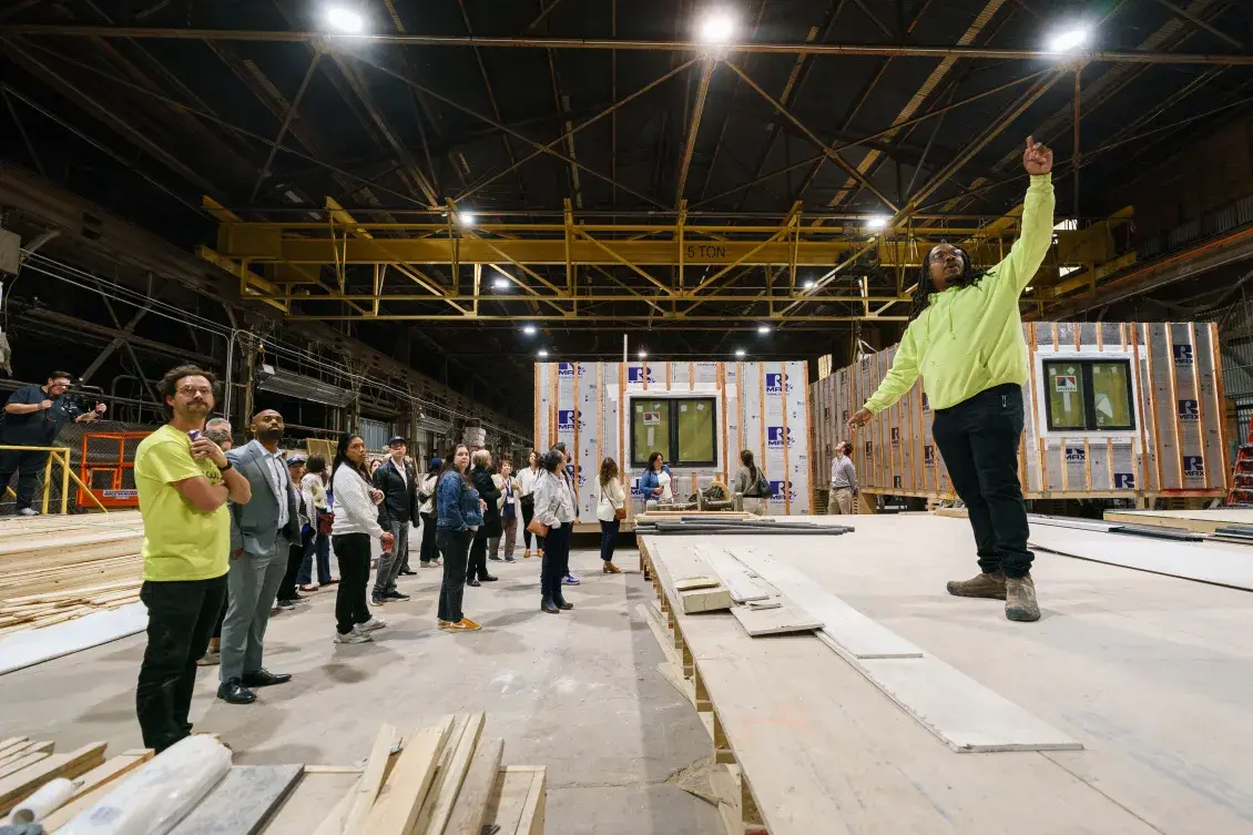 Person in neon yellow sweatshirt stands inside modular housing factory speaking to a group of people. There are modular structures in the background.