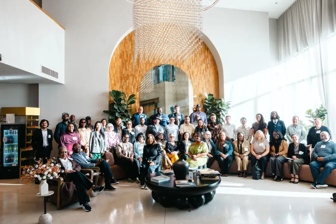 A group of people pose together in a sunlit room with high ceiling in front of a white wall with a tan wooden arch panel