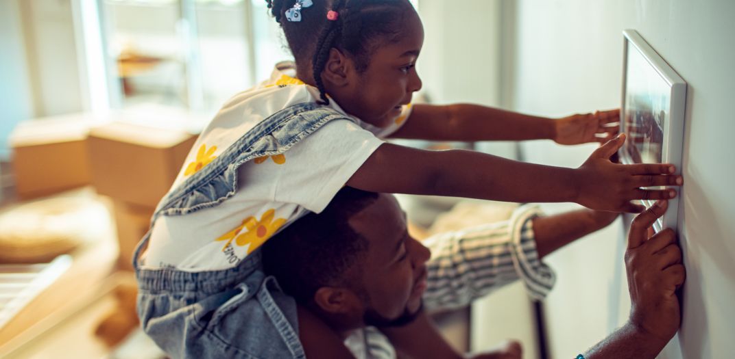 A girl sits atop a man's shoulders as they hang a photo on the wall