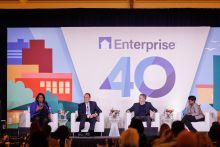 A group of panelists sit in white arm chairs on a stage featuring a large, colorful backdrop.