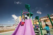 Child stands at the top of the slides and watches another child slide down. A third child runs by the slides.