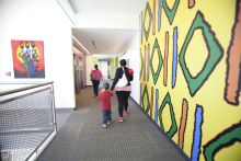 Children and adults walk down a hallway by a yellow wall painted with multi-colored shapes