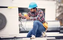Person in plaid shirt wearing a blue construction hat kneels down to work on a heat pump.