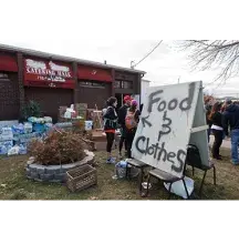 A group of people stand by boxes in front of a building with a red awning that reads Catering Hall and a sign that reads Food and Clothes 