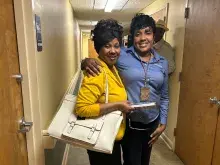 Two people stand next to each in a hallway, with the person on the right wearing a blue shirt and wrapping her arm around the shoulder of the person in a yellow shirt who carries a large white tote purse.