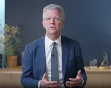 Person with short silver hair wearing blue suit jacket, blue tie and white dress shirt speaks and gestures with hands. A table with a plant and a blue wall are in the background.