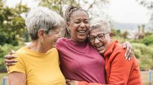 Three older women hug and laugh in front of green space.