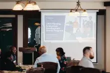 A group of people sit at a roundtable in front of a screen that reads "USVI Housing Ecosystem Development Grants Program"