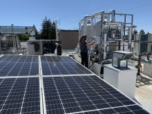 Rooftop view of flat solar panels with a worker adjusting a heat pump; blue sky and trees are in the background