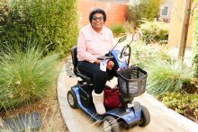 A woman on a scooter she uses as a mobility aid smiles from an apartment community's leafy green space.