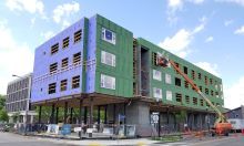 Corner multifamily apartment building undergoing a window installation; a crane is shown on the right with a blue sky and a few clouds in the background