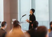 Person stands at a podium speaking into a mic before an audience of people