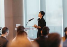 Person stands at a podium speaking into a mic before an audience of people