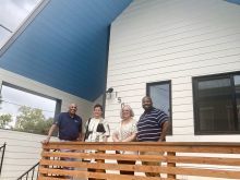 Four people standing on the porch of a white house with blue awning