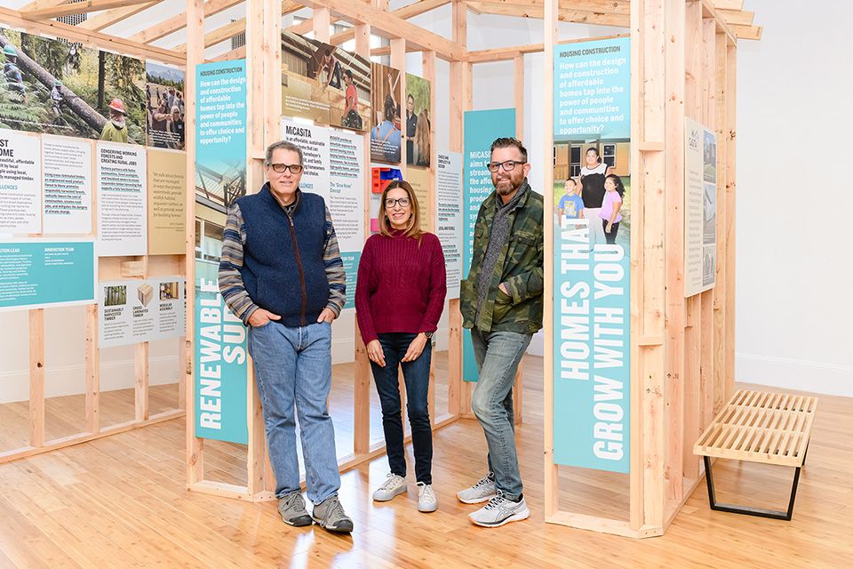 A woman and two men standing in front of a wooden structure exhibit at the Enterprise A Better Way Home Exhibition