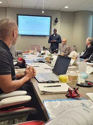 A presenter speaks to meeting participants sitting at a conference table and taking notes with notebooks and laptops