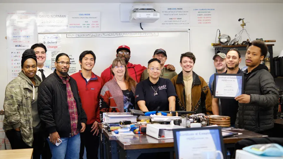 Group of people pose in a classroom with a whiteboard in the background; one person on the right holds up a certificate.