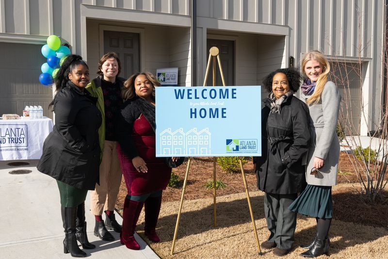 5 woman in winter jackets standing outside near a sign that says welcome home.