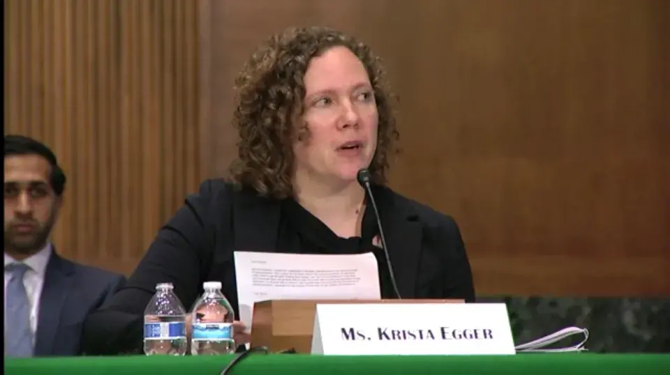 Person with medium length hair and a black blazer seated and speaking at a table with wood paneling in the background.