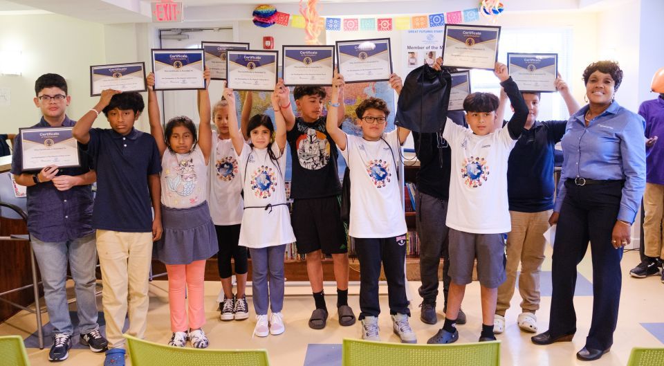 A teacher stands with a group of children stand in a line holding their certificates high in a classroom