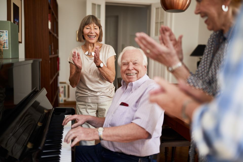 An older adult plays the piano as friends watch on and clap