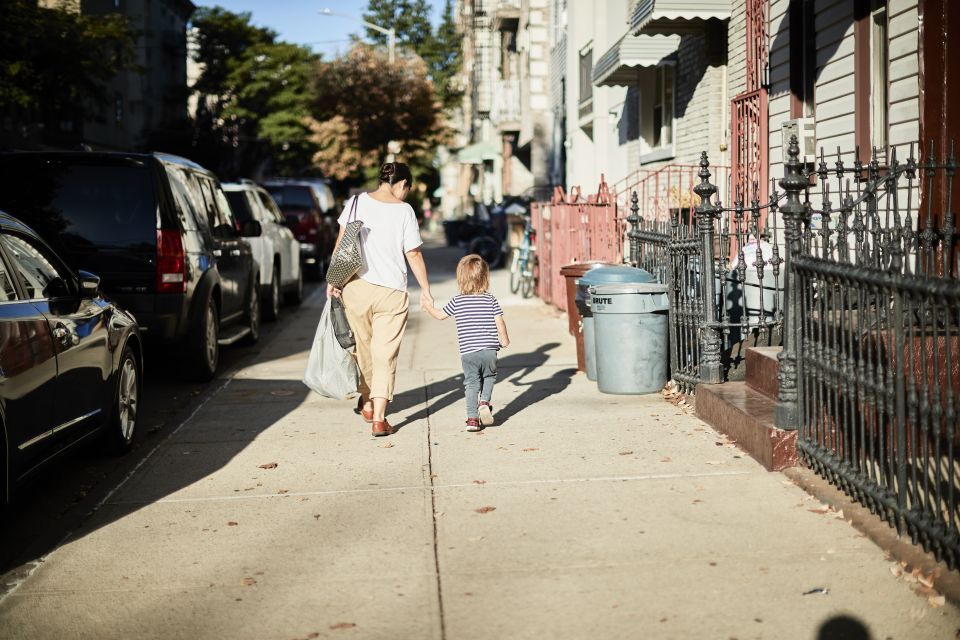 A mother and child hold hands as they walk on a sidewalk alongside buildings and cars parked on the street