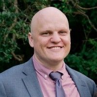 Person with shaved hair wearing a maroon dress shirt and a blue tie and blazer