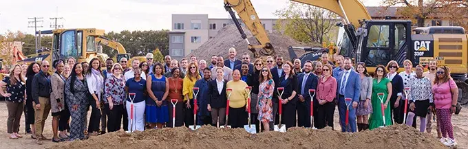 A large group of people standing at a construction site with construction equipment. The first row of people is holding shovels.