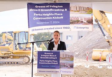 Janine Lind, president of Enterprise Community Development, stands at a podium for a groundbreaking ceremony with construction equipment in the background