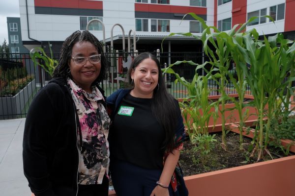 Two smiling women standing in front of plants with apartment building in background