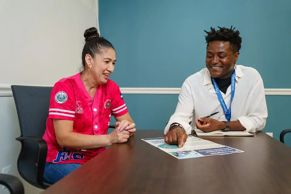Person with a top bun and bright pink top sits at a table with a person with black hair wearing a white shirt, pointing at a flyer
