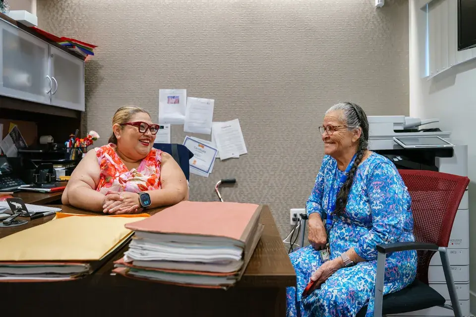 Person with glasses wearing brightly colored top sits at a desk next to person with a long gray braid wearing a blue dress, both smiling