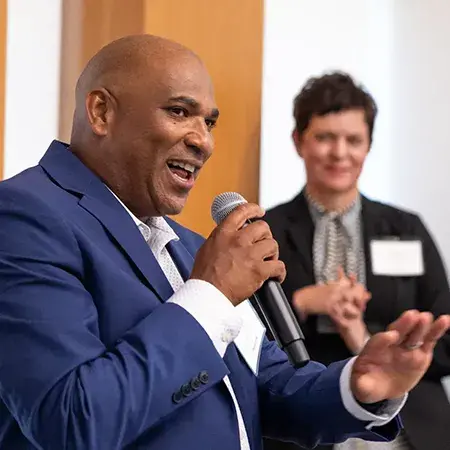 Oji Alexander speaks into a microphone with Michelle Whetten, VP and Market Leader for the Gulf Coast behind him