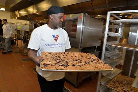 A man holds a large tray of baked goods to place in a rack of a commercial kitchen