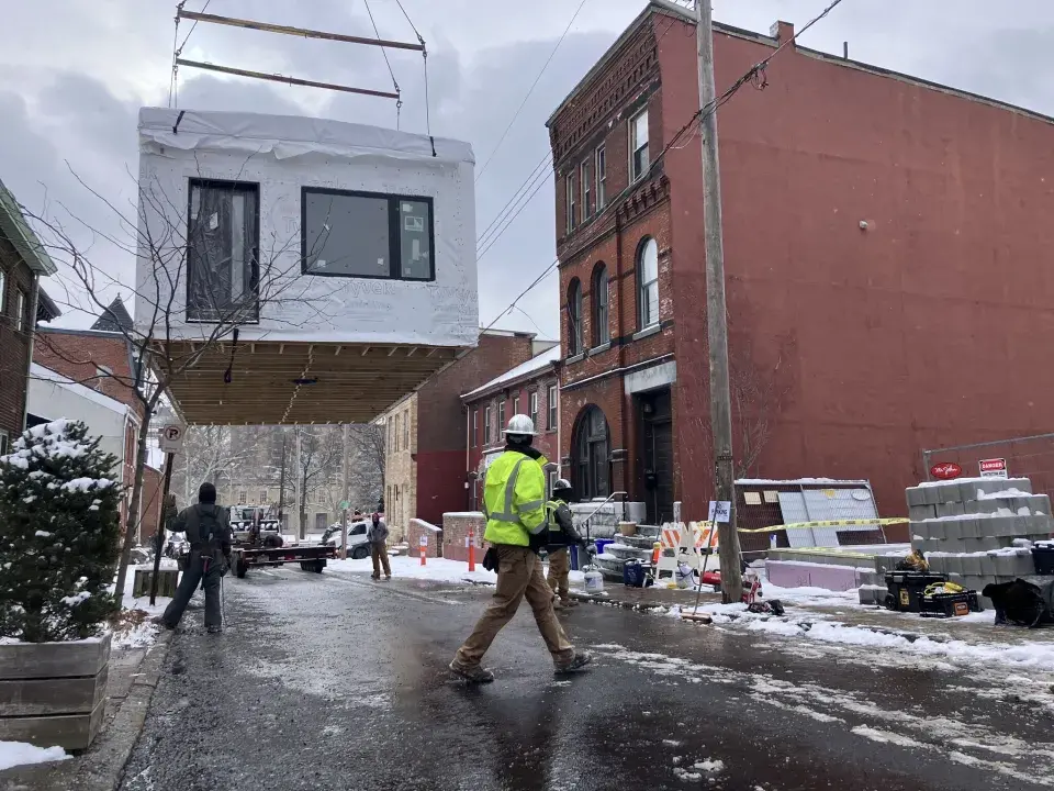 A modular house being carried by a crane as a construction worker stands in front, with gray sky and slush on the street