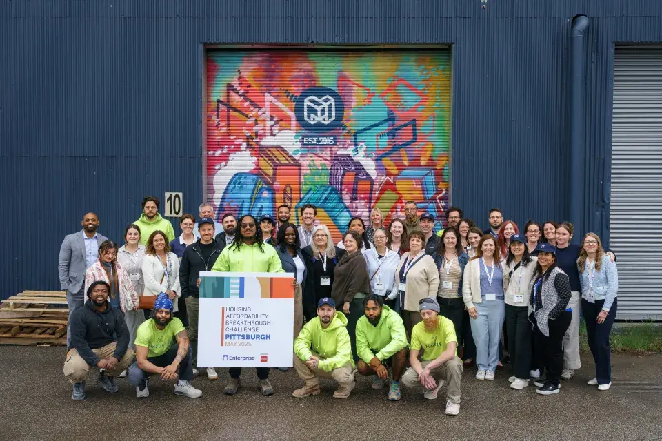 Group of people stand in front of the outside of a industrial building with a colorful mural. A sign reads: Housing Affordability Breakthrough Challenge