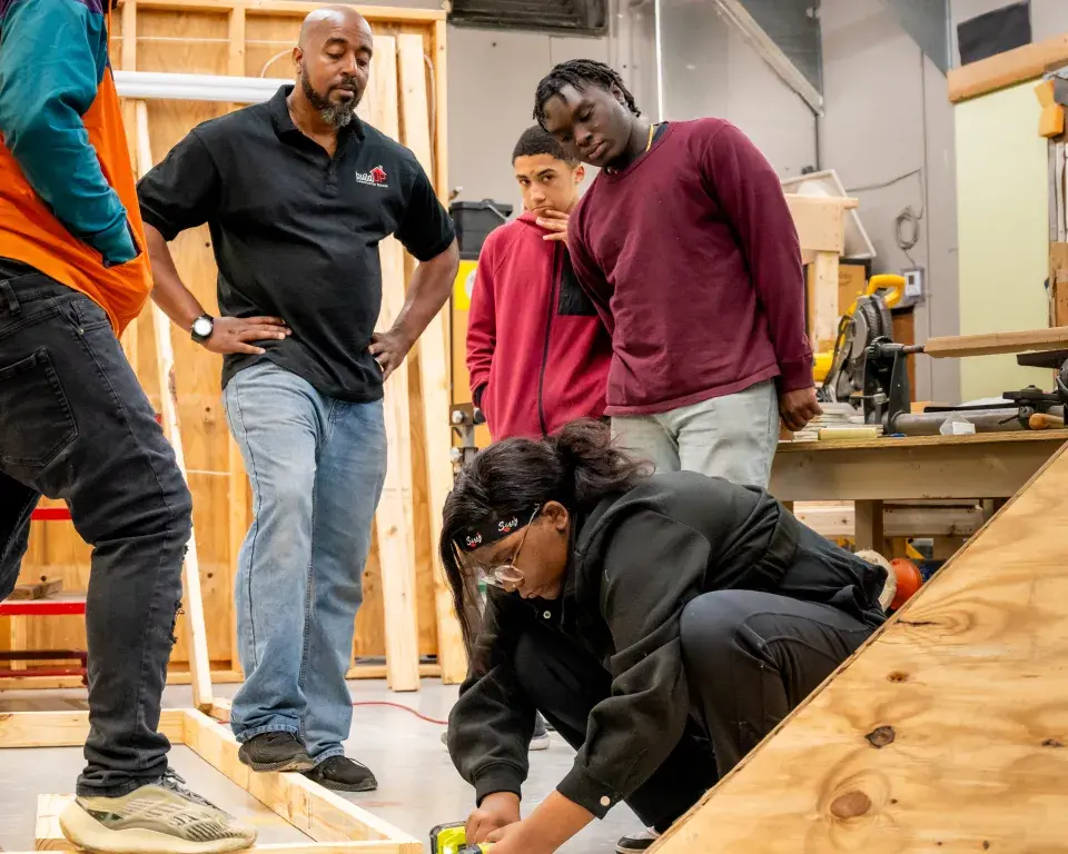 Young person measures construction material while teacher and students observe